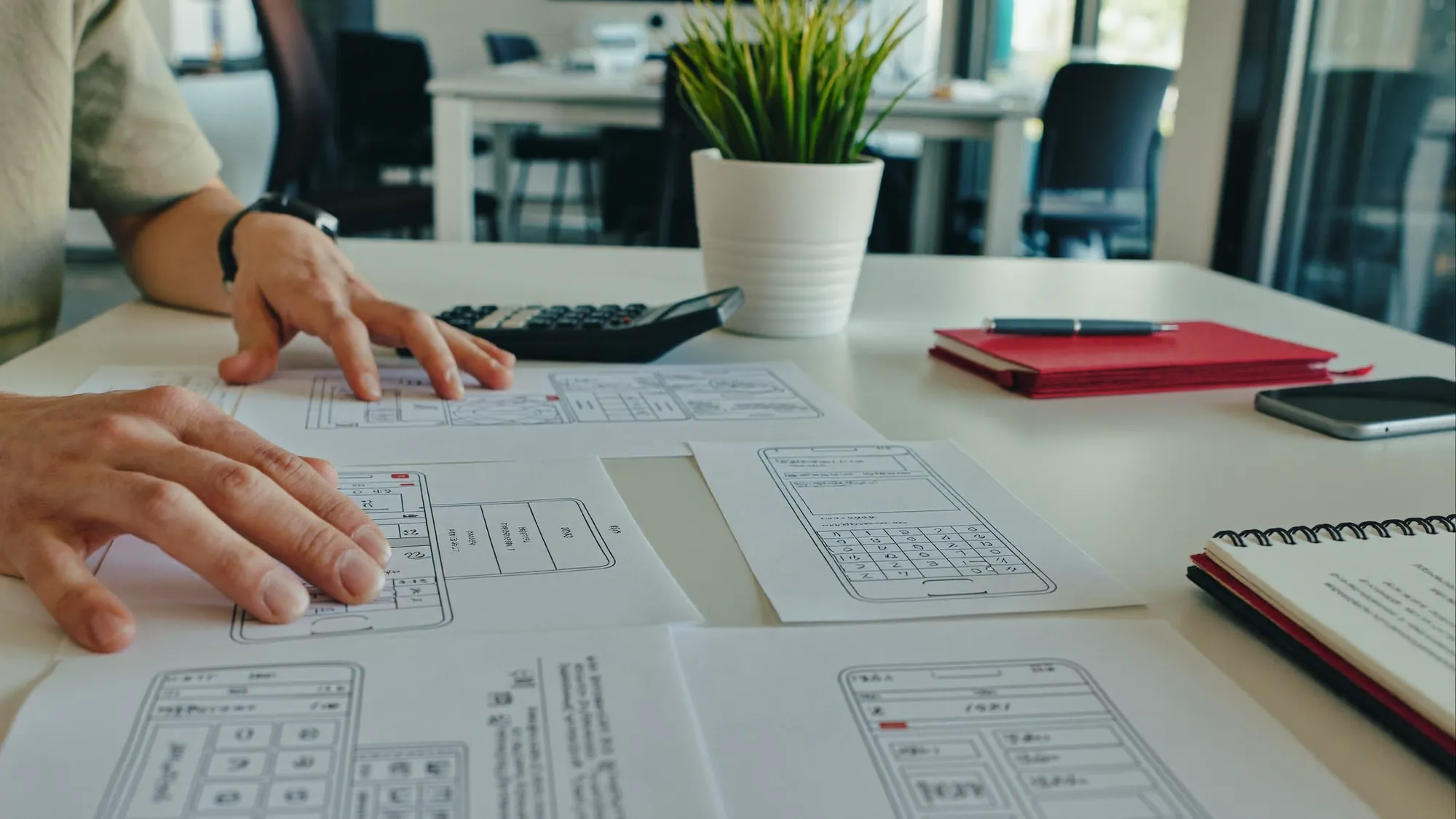 Manos revisando bocetos de wireframes de una app móvil con una calculadora, ilustrando el costo de desarrollo de una app móvil